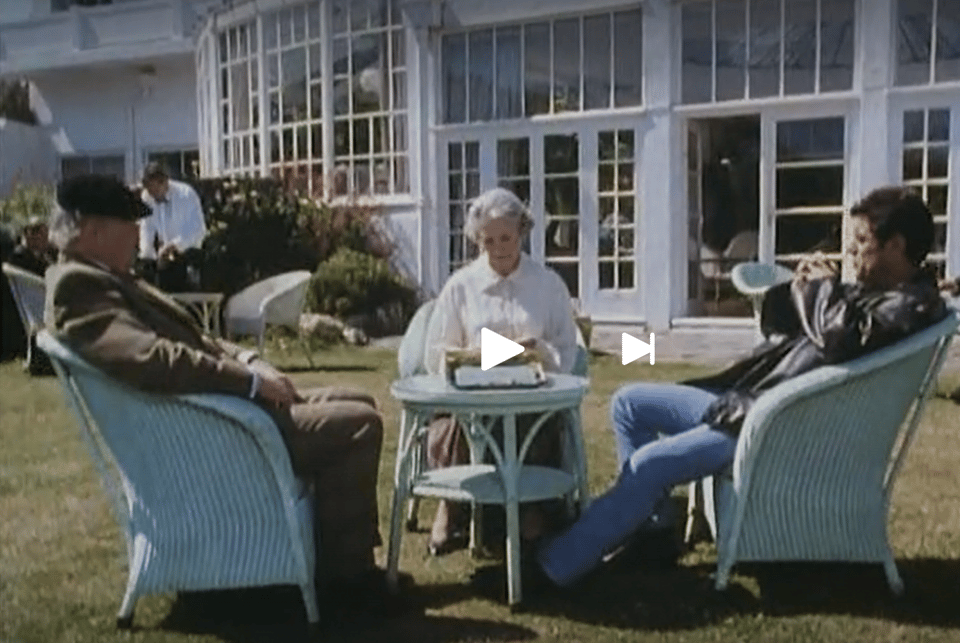 Screenshot of three characters sitting in Lloyd Loom chairs on the lawn of Burgh Island Hotel. The restored palm court is behind them.