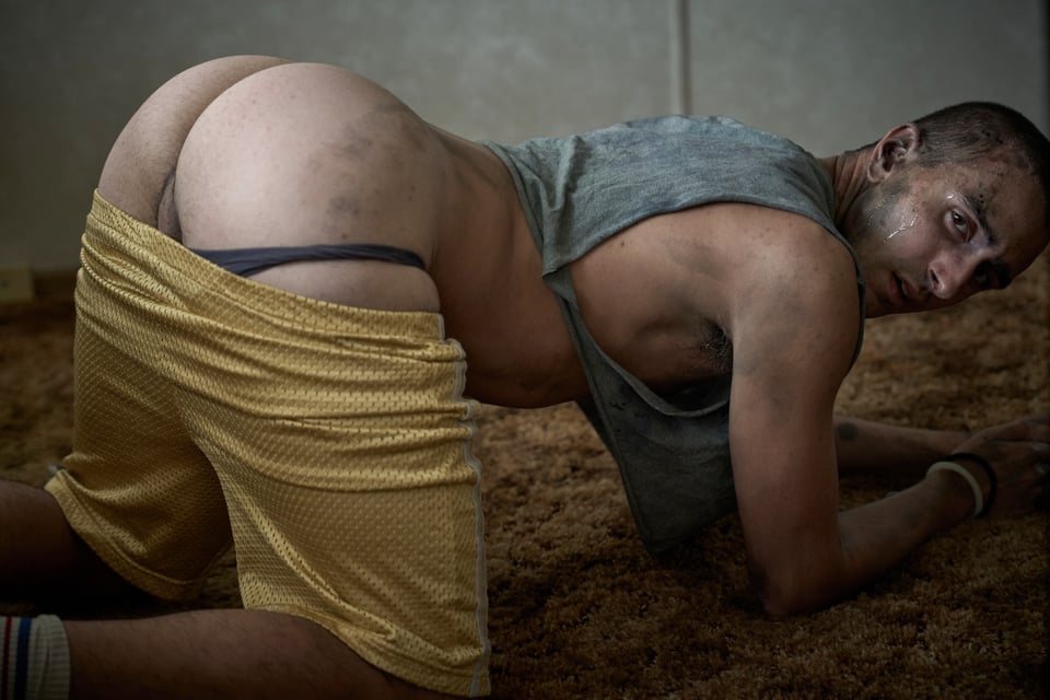 shaved head man kneels on all fours with his gym shorts pulled down