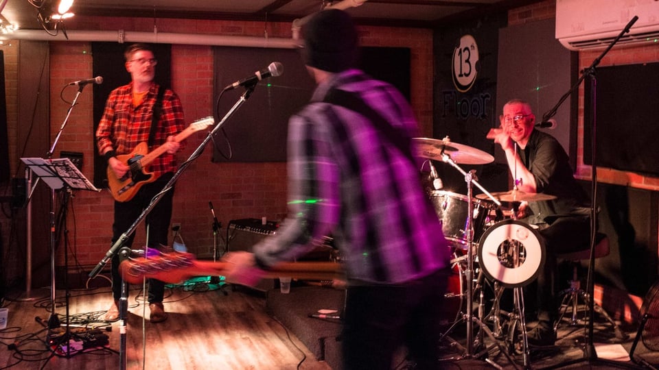 A man playing guitar looks to his left at a man playing drums, who is looking at a man playing bass in the foreground, with his back to the camera.