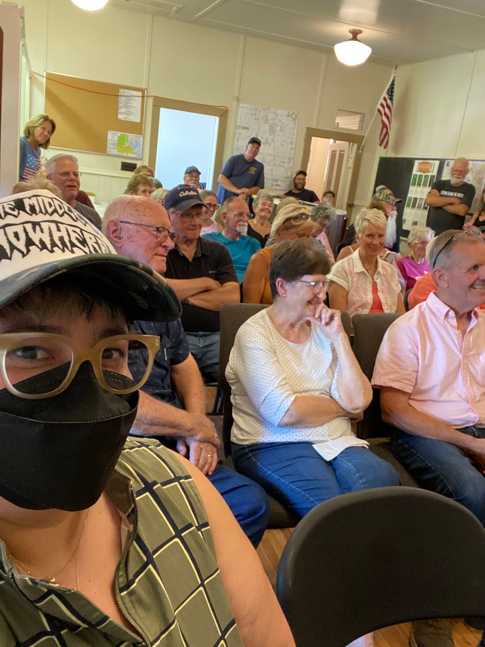 a selfie with a queer chicanx person wearing a kn94 mask foregrounded in front of a crowd of mostly elderly white people