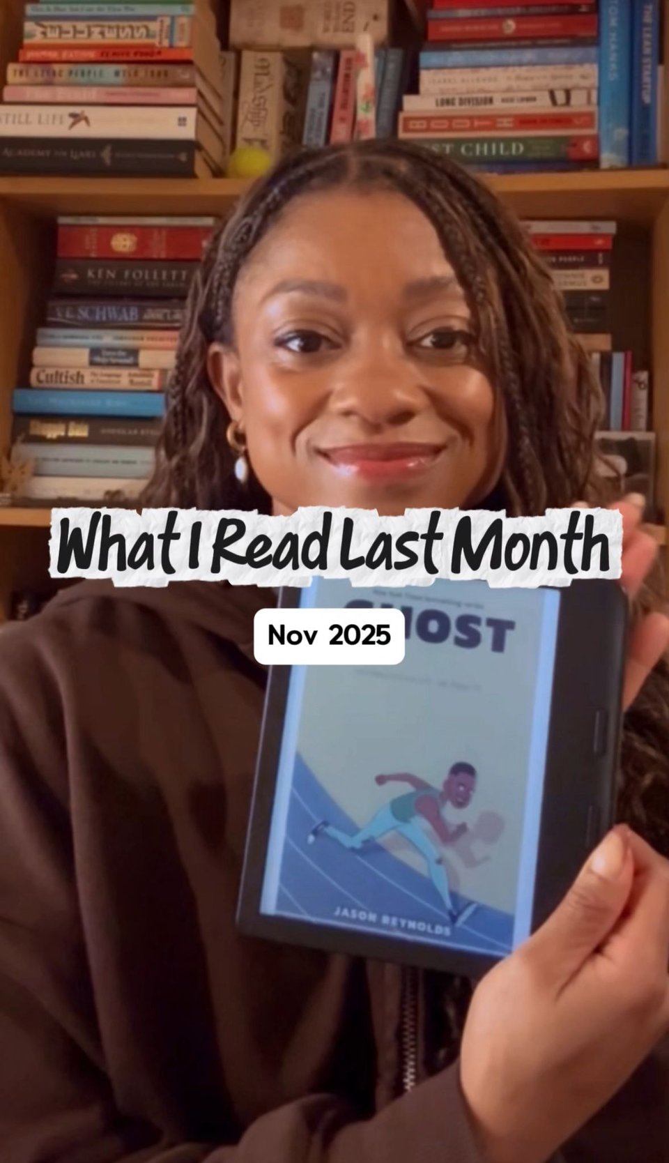 A woman smiling and holding an e-reader in front of a bookshelf. Overlaid text reads “What I Read Last Month” with a label underneath saying “Nov 2025.” The e-reader displays a book cover.