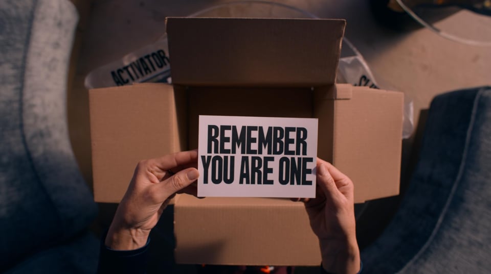 Hands hold a card that reads "Remember You Are One" over an empty cardboard box.