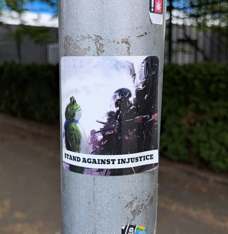 A close-up photo of a signpost with a sticker on it. In the sticker, a large green frog faces off with ICE thugs in riot gear. The sticker is captioned "Stand against injustice." We side with the frog.