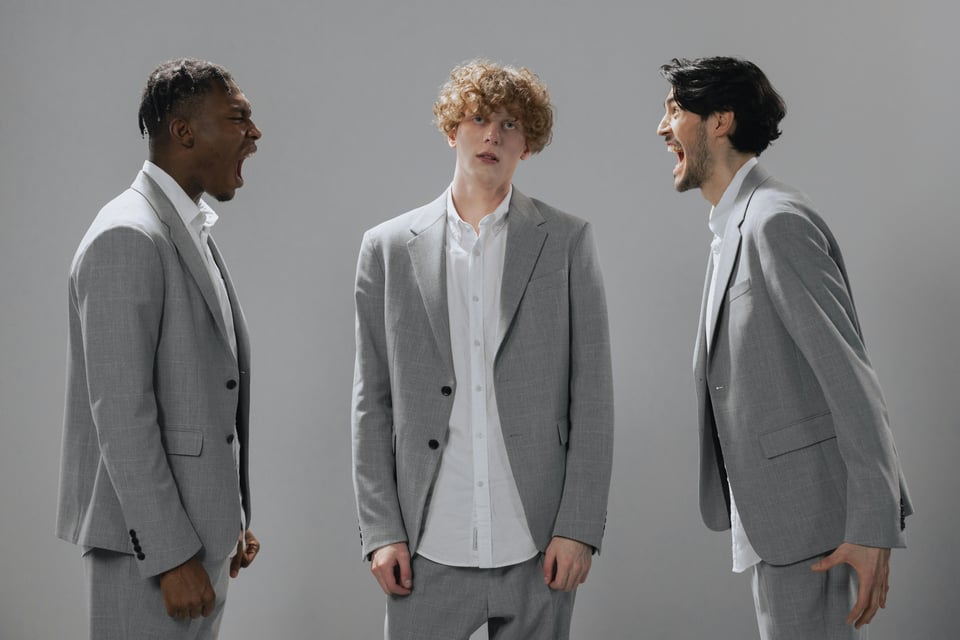 Three men in light gray suits stand in a studio setting. The man in the center has curly hair and a neutral expression, while the two men on either side are facing him and yelling animatedly. The background is plain gray.