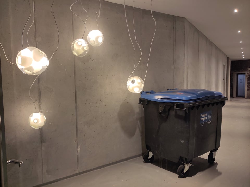 Blick in einen mit Waschbeton ausgekleideten Gang. Darin steht eine Mülltonne, neben ungeordnet von der Decke hängenden, runden, etwa fußballgroßen Lampen.