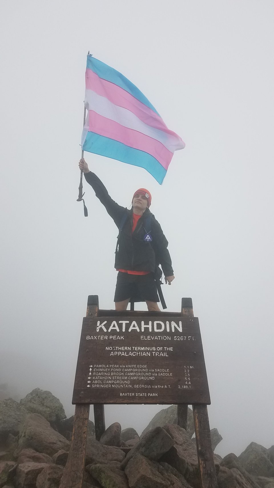 A picture of me in 2021 standing on the summit of Katahdin in Maine, the northern terminus of the Appalachian Trail. I wave a trans pride flag above me.