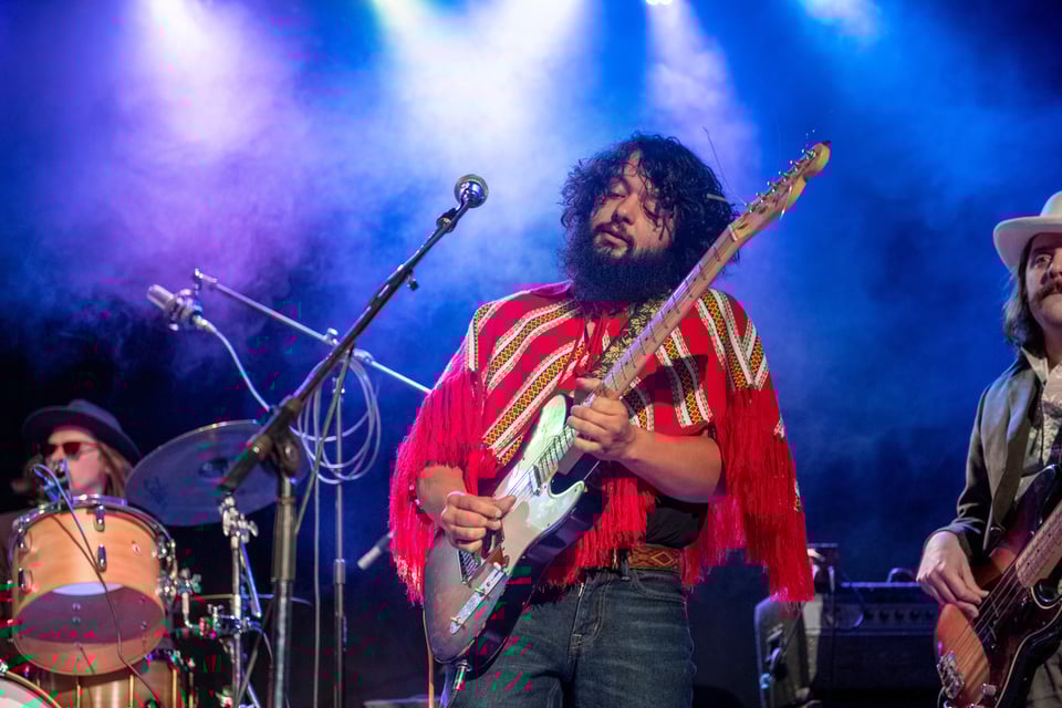 A man with a dark, bushy beard wearing a red sarape plays a Fender Telecaster guitar.