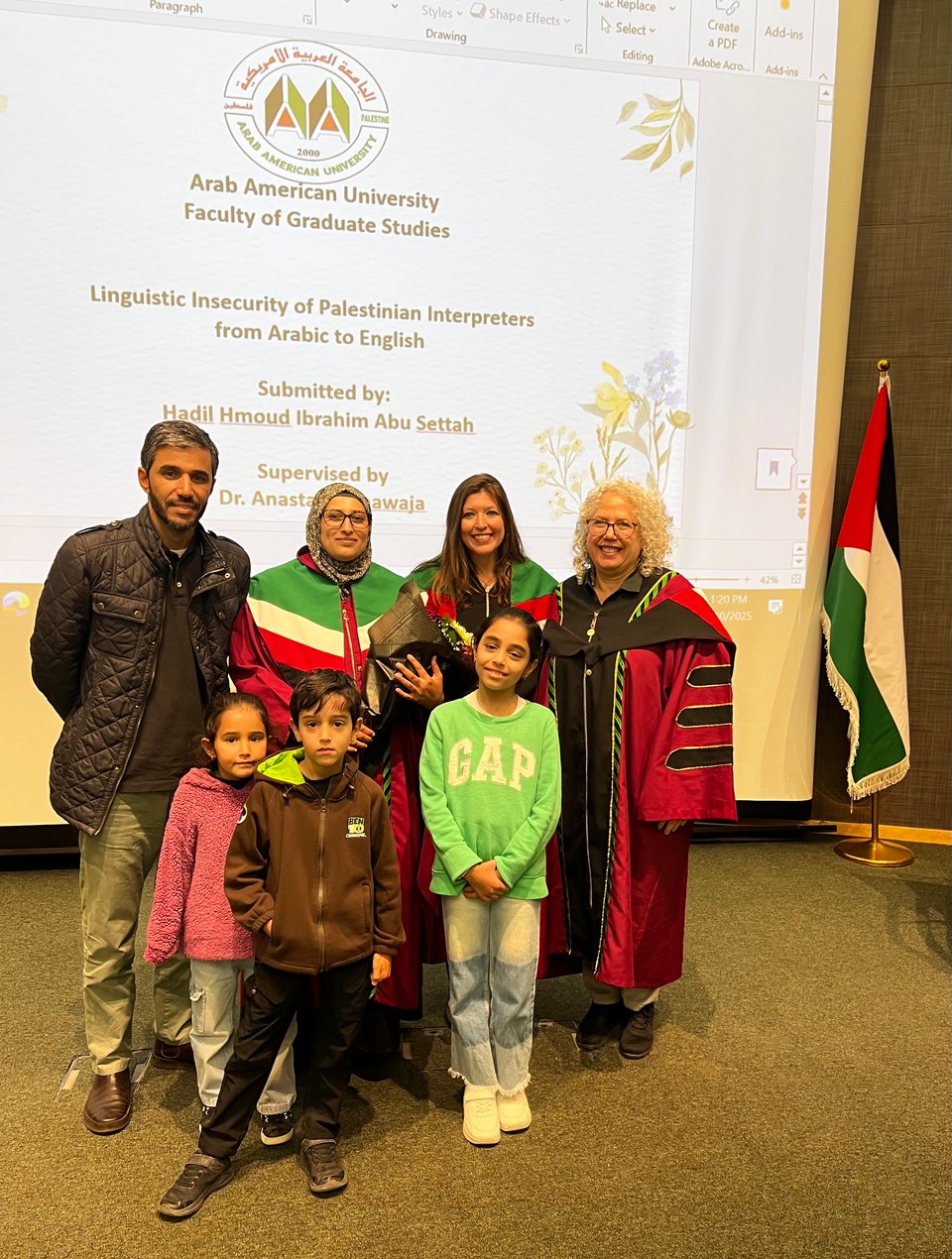 7 people standing in front of a large screen; four adults (one man and three women) and three children standing in front of the adults. A Palestinian flag is in the background
