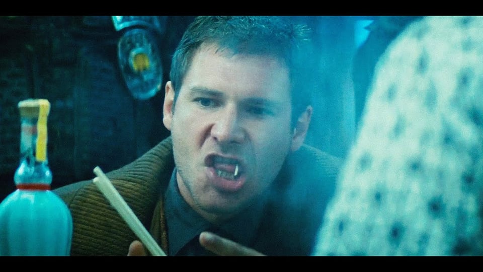 an image of harrison ford playing the character rick deckard in blade runner. he is sat at a noodle bar with a single noodle hanging out of his mouth.