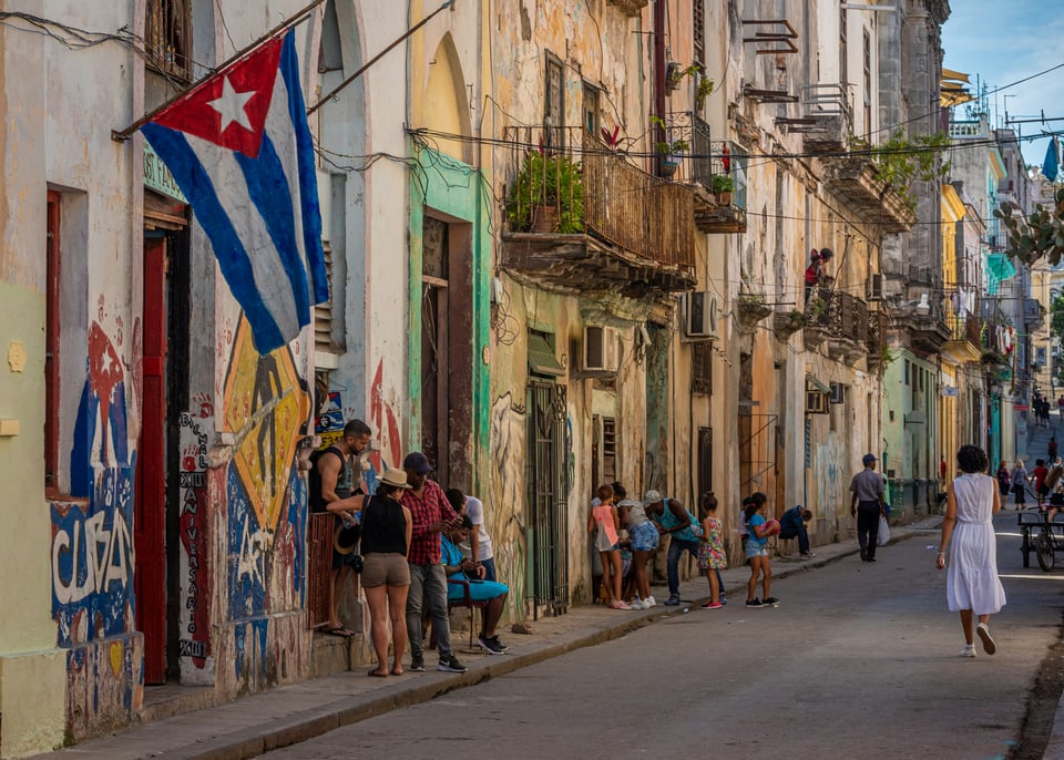 People gather along the street in front of buildings in Havana, Cuba. A Cuban flag hangs from the building on the left side. Graffiti on the walk below the flag has a picture of the Cuban flag and Cuba painted on the wall, alongside a pedestrian crossing warning sign.