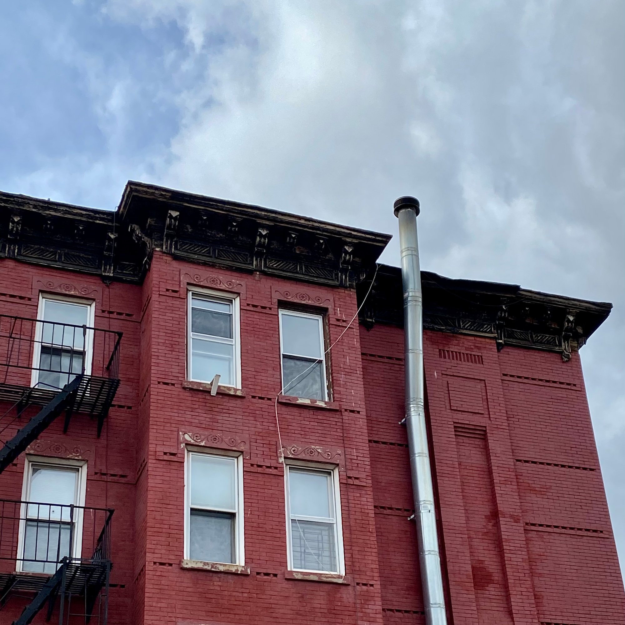 Image of cloudy sky and a red brick building with a black iron fire escape on the left and a long, silver pipe on the right conversing with a loose string hanging from the roof down to the third floor window. Where is it going?