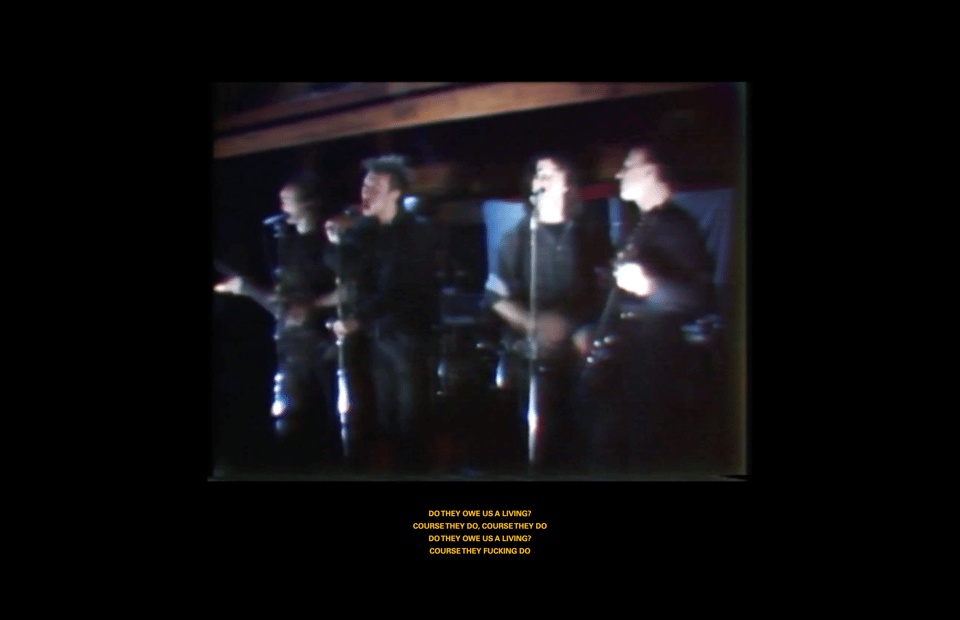 A screenshot of a presentation slide: A video of Crass performing is central, with subtitles displaying the chorus of 'Do They Owe Us A Living?'