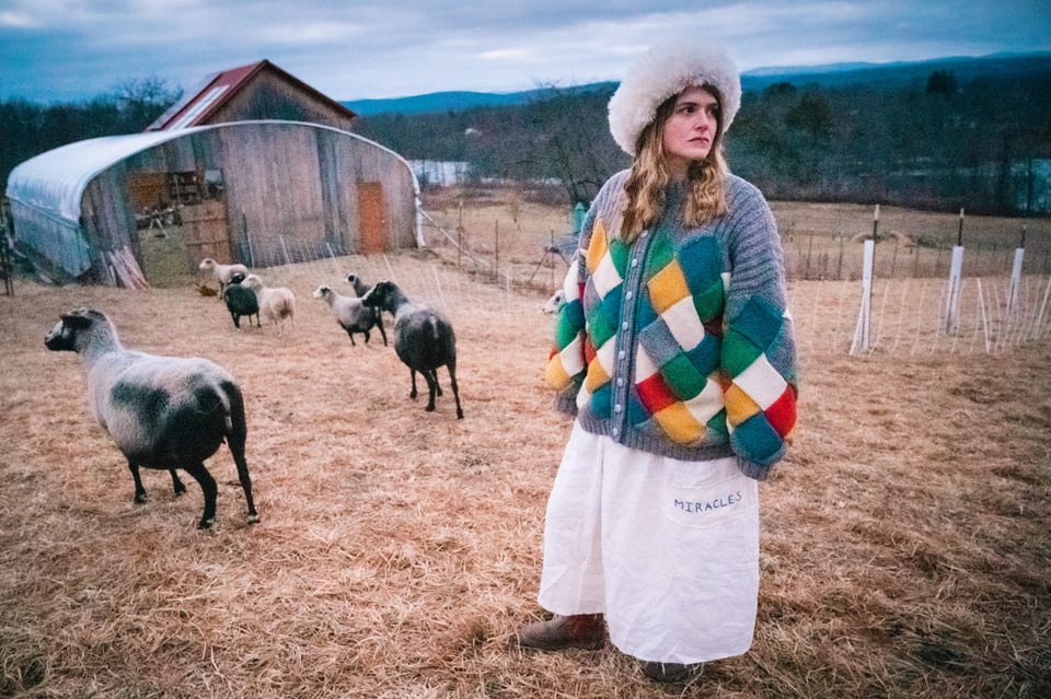 A woman with blond hair past her shoulders stands in a sheep enclosure on a cloudy day. She wears wide-legged white pants, a multi-colored quilted patchwork and a big furry white hat. There are a number of sheep in the background, near a low structure.