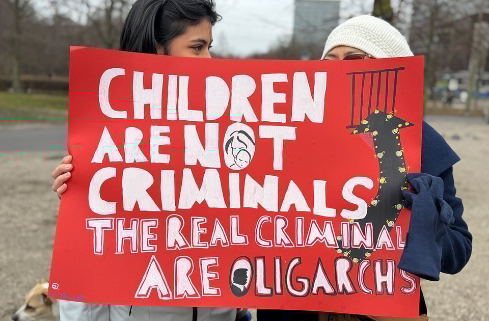 Two women hold up a sign that says "Children are not criminals. The real criminals are oligarchs".