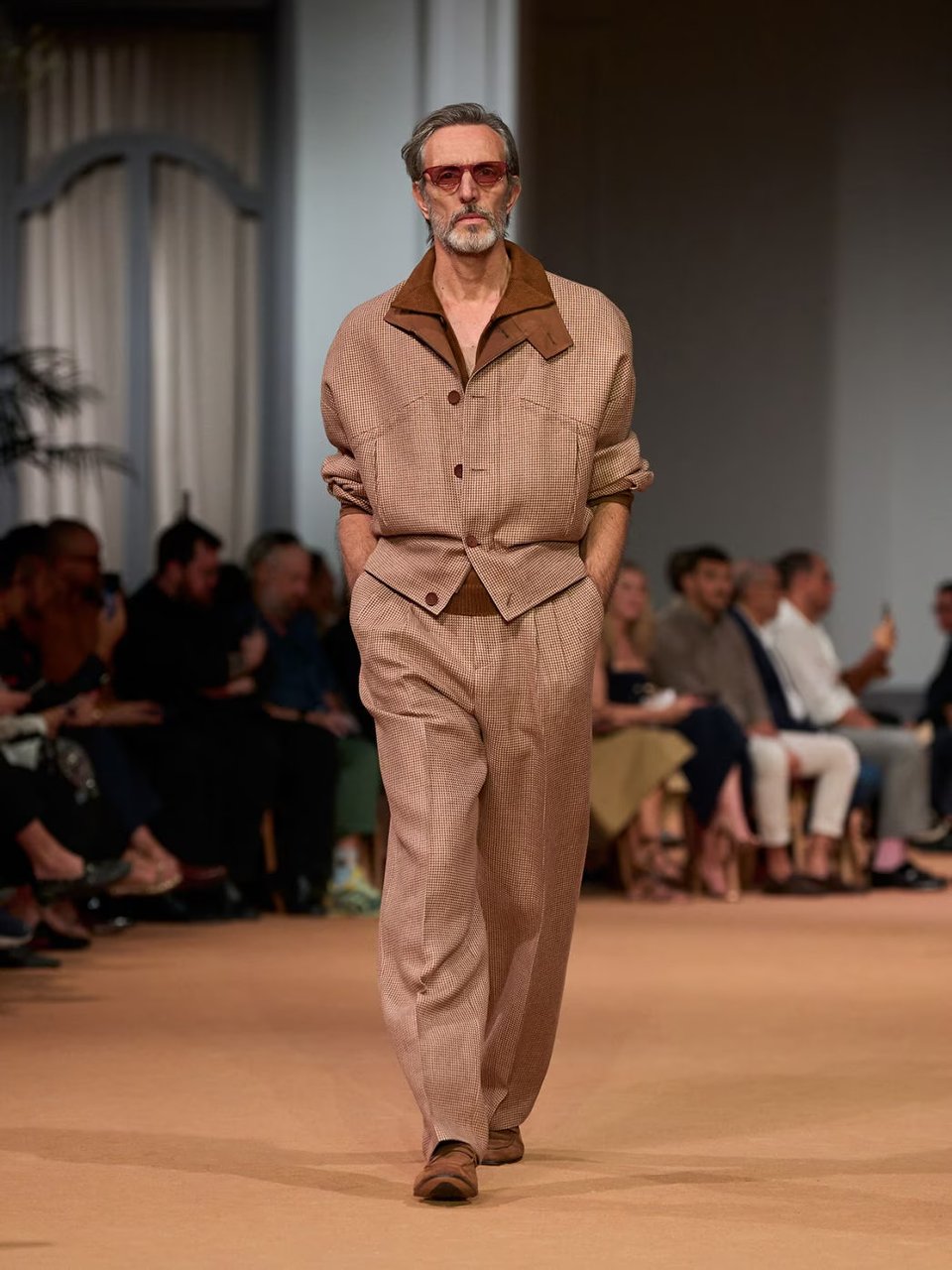 Photo of older white male model walking in a Zegna fashion show, wearing a tan leisure suit with a brown shirt