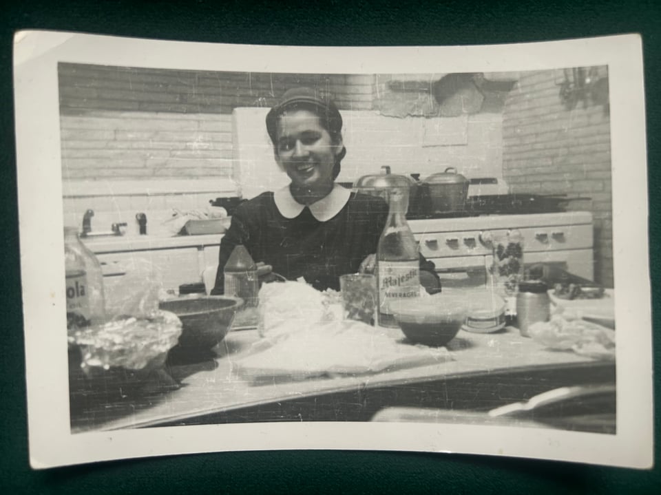 a black and white photograph of a fair-skinned Black woman sitting at a kitchen table with a white stove in the background.
