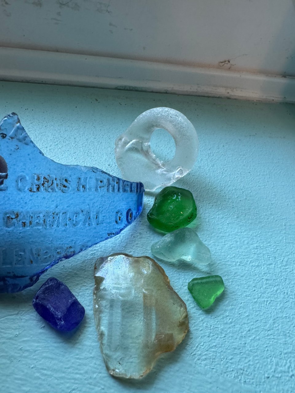 Bits of blue, green, teal, and clear river glass on a windowsill. Once piece contains the letters TL. Another has a chemical notice imprinted in the glass. There's also a small cup or jug hangle.
