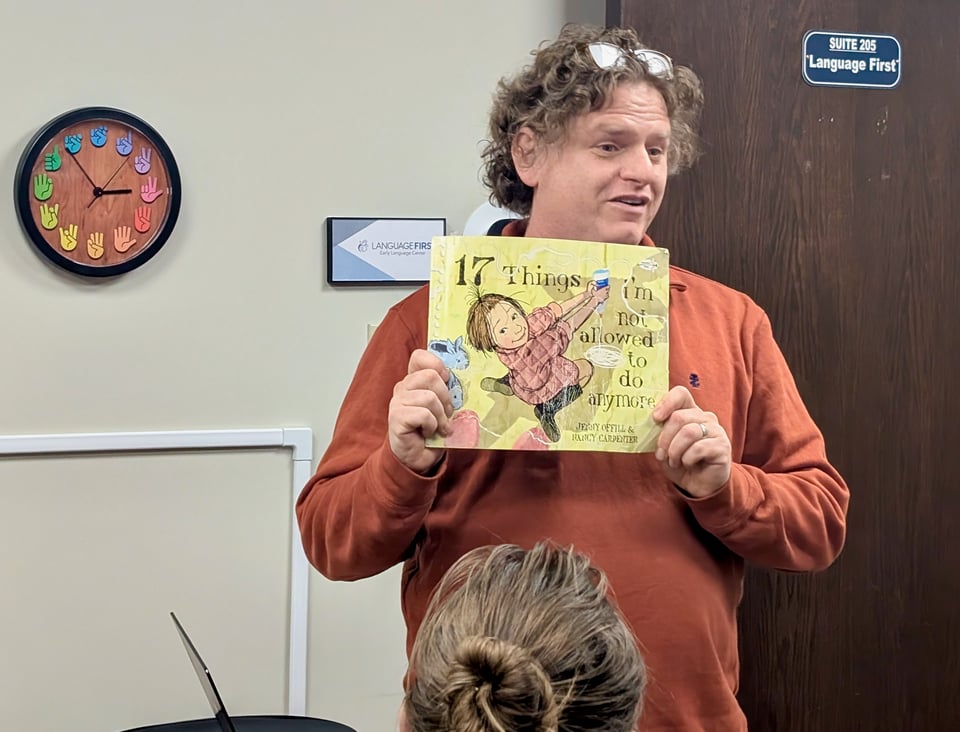 A middle-aged white guy, Will Fertman, stands in a small office space wearing a rust-color sweater. He's giving a talk, holding up a children's book titled "17 Things I'm Not Allowed to Do Anymore"