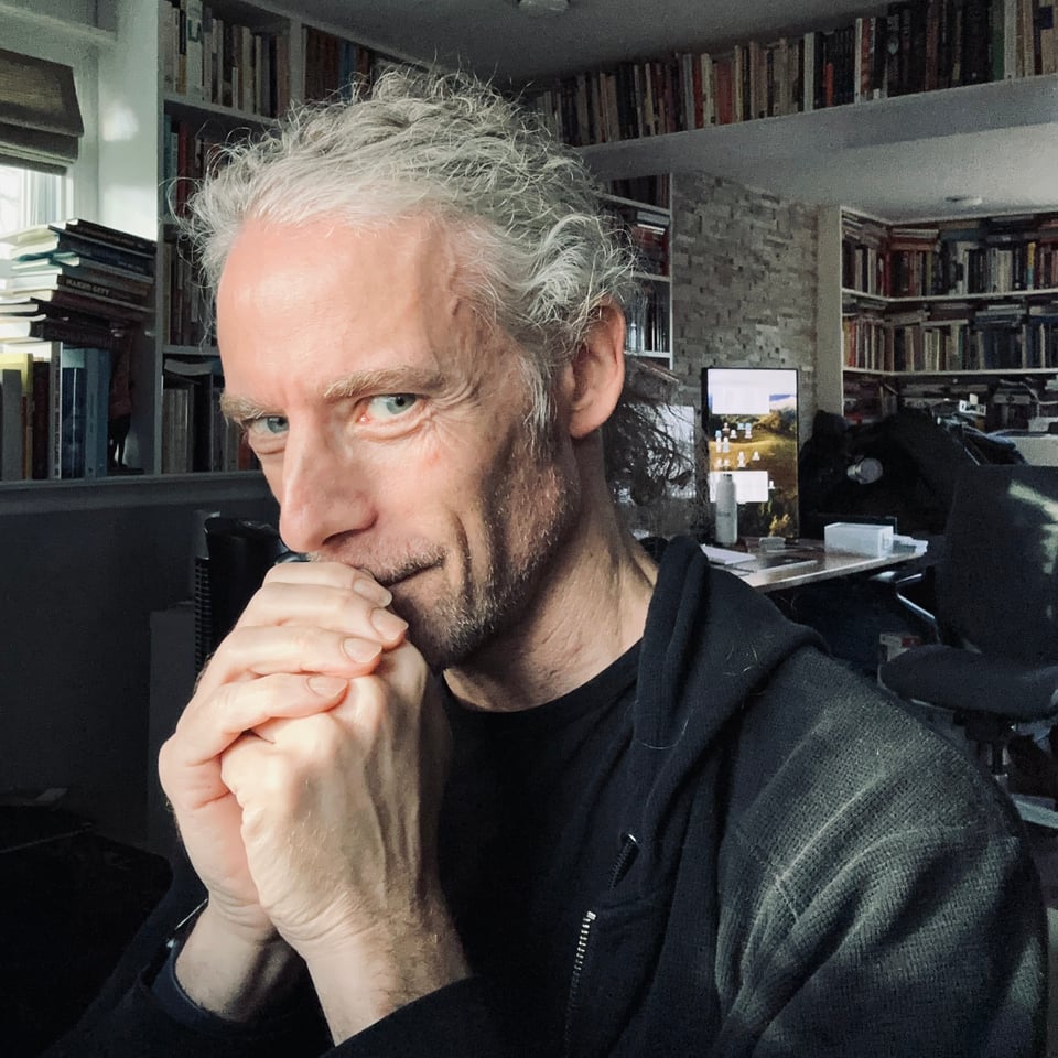 Selfie of white man with scraggly hair tied back, smiling tight-lipped at the camera in 3/4s profile, wearing a black T-shirt and black zip-up hoodie. There are books on shelves behind him