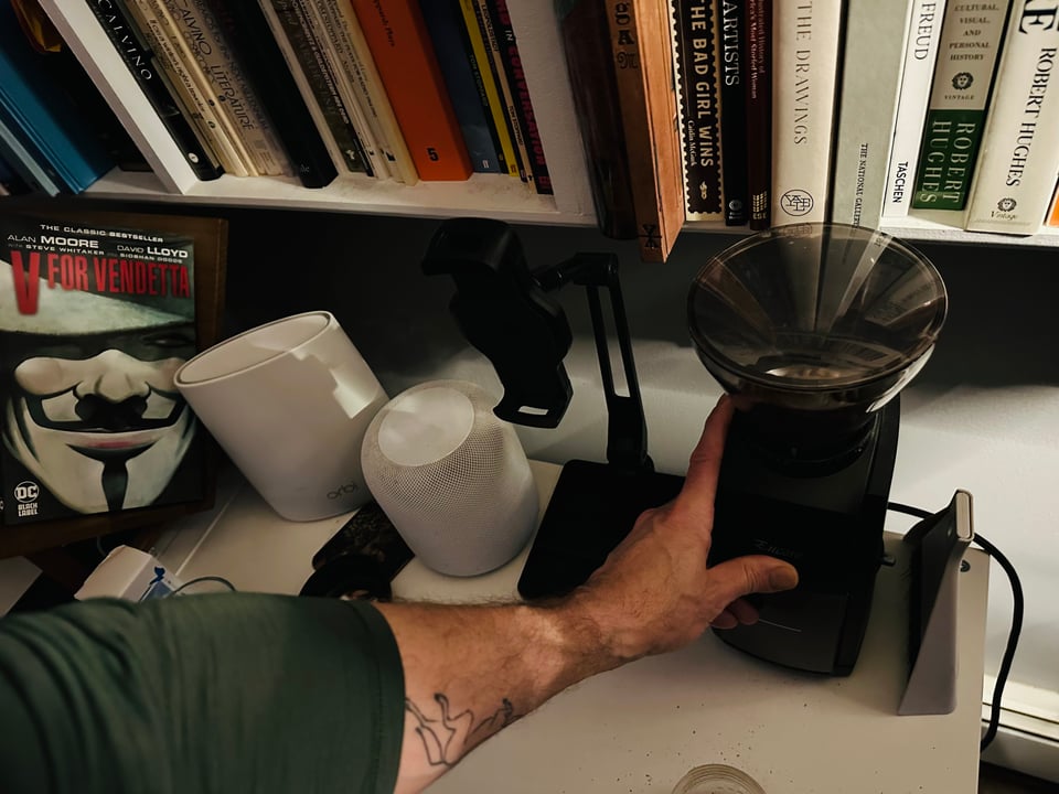 Photo of white man’s arm reaching down to start coffee grinder; we can see a bookshelf above the grinder, and to the left, an Apple HomePod (white), a wifi router, and the cover of Alan Moore & David Lloyd’s V for Vendetta