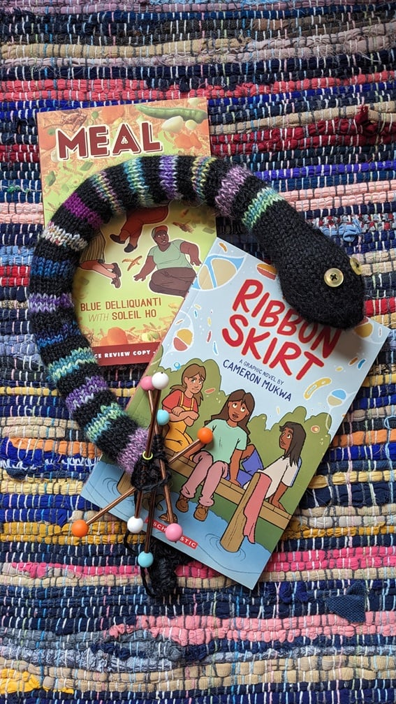 a photo of an in-progress knitted snake laying on top of two graphic novels that Maia had just finished reading: Meal by Blue Delliquanti & Soleil Ho and Ribbon Skirt by Cameron Mukwa. The snake and books are laying on top of a brightly colored rag rug. The snake has 15 stripes, representing 15 books read so far at the time the photo was taken.