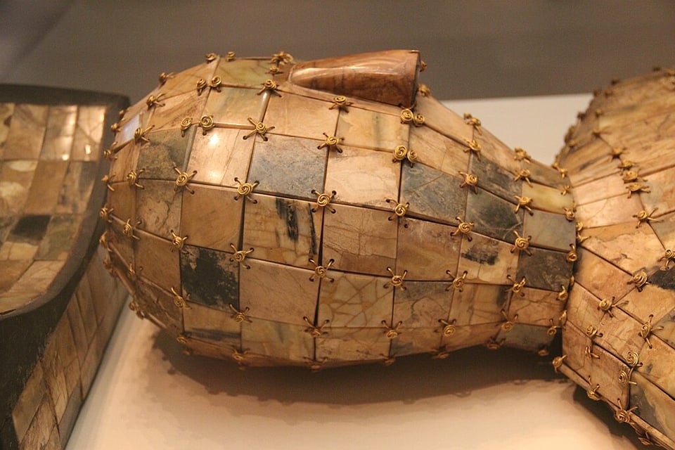 A jade burial suit, sandy colored with golden thread. It looks like several squarish tiles fastened together in the shape of a person. The image focuses on the head, but the torso can be seen.