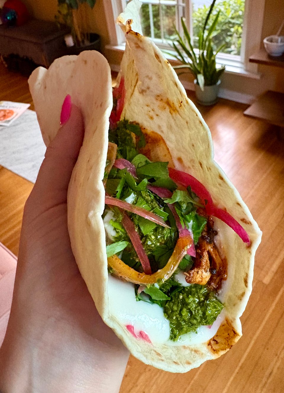 The author's hand holding a Navajo flour tortilla stuffed with Arabic-spiced al pastor chicken, toum, green shatter, herbs, and pickled red onion.