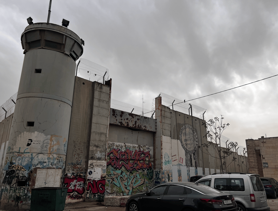 A concrete barrier wiht barbed wire at the top, and a military tower with 360 degree windows at the top. The wall is part of a parking lot, and has graffiti painted on it 