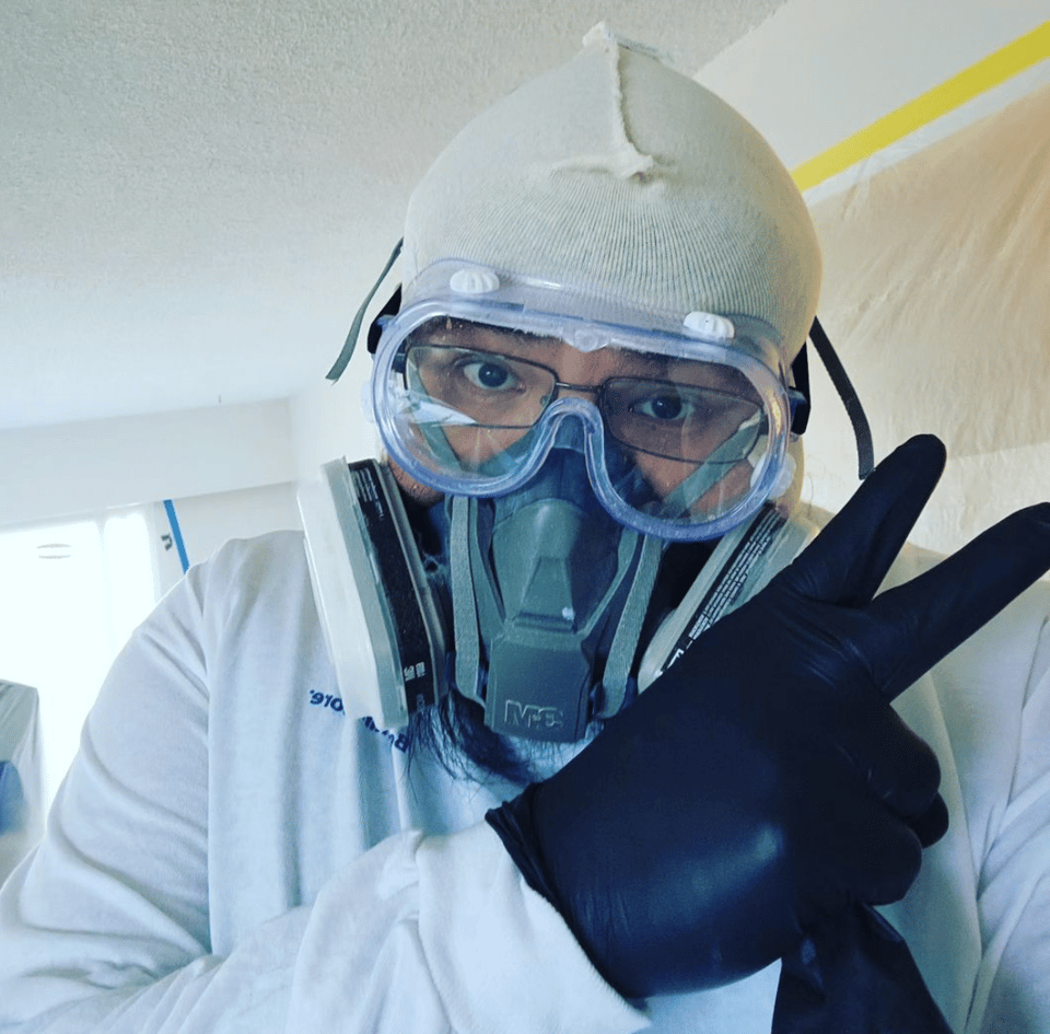 A Man wearing a white cap to protect his hair, goggles, breating protection, a white jacket, and black gloves.