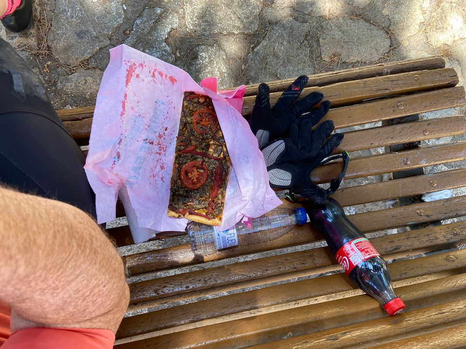 a slice of pizza on a bench, the pizza features tomatoes and some sort of vegetable. next to it is a bottle of coke, a too small empty bottle of water, some gloves and a pair of sunglasses