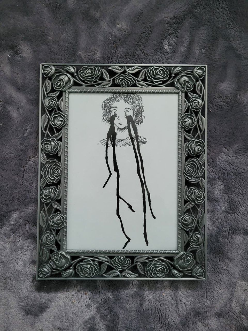 Photo of a metal rose frame containing a black and white drawing of a person crying. The tears drip long and take up most of the length of the drawing
