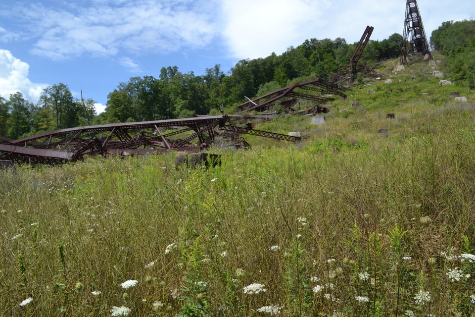 Part of a collapsed iron bridge tumbles down a fairly steep slope, a nearly intact tower can be seen at the top. The ground is covered in tall grass and wildflowers.