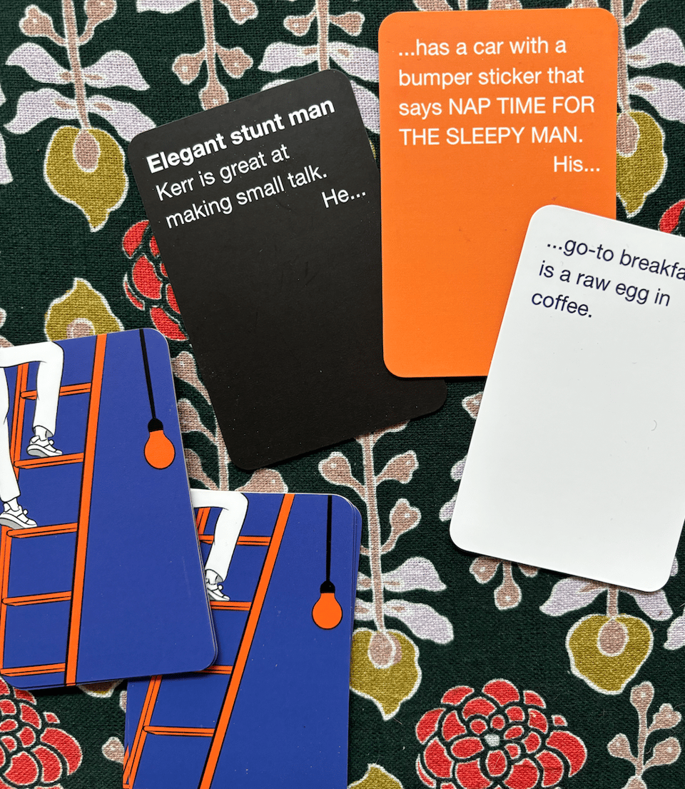 A few cards dealt out onto a patterned background. Three of them are visible: they read "Elegant stunt man Kerr is great at making small talk. He..." "Has a car with a bumper sticker that says NAP TIME FOR THE SLEEPY MAN. His..." "go-to breakfast is a raw egg in coffee."