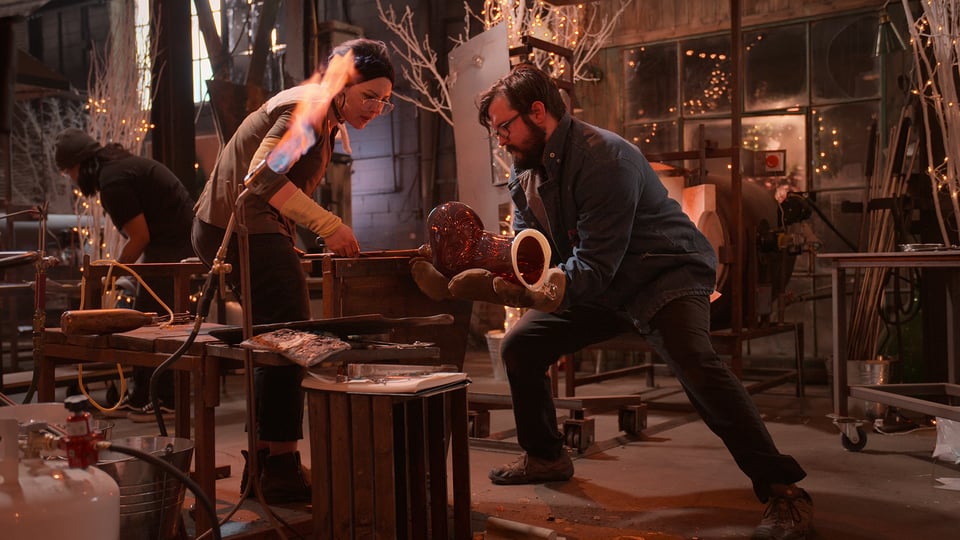 a man and woman working on a red boot-shaped 3D glass object a workshop decorated for the holidays