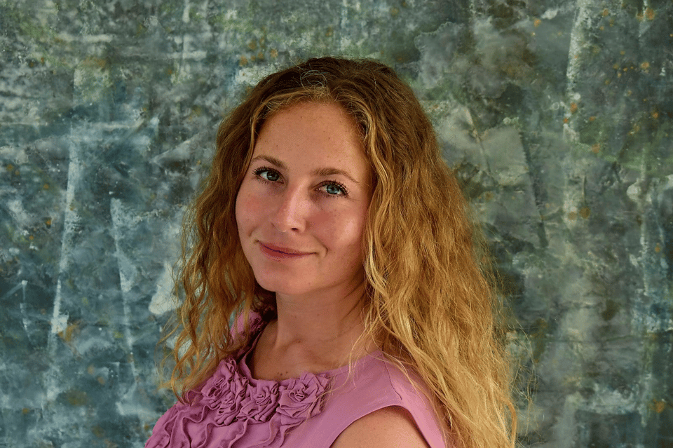 Picture of Amber van Veghel, a white woman with blonde curly hair and a pink flowery outfit, matching with a green stone like background.