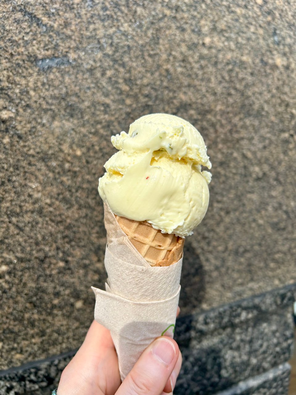 A white person's hand is holding an ice cream cone wrapped in a beige paper napkin. the cone is topped with rich golden ice cream with tiny flecks of pink and green. Image by Rowan Ambrose.