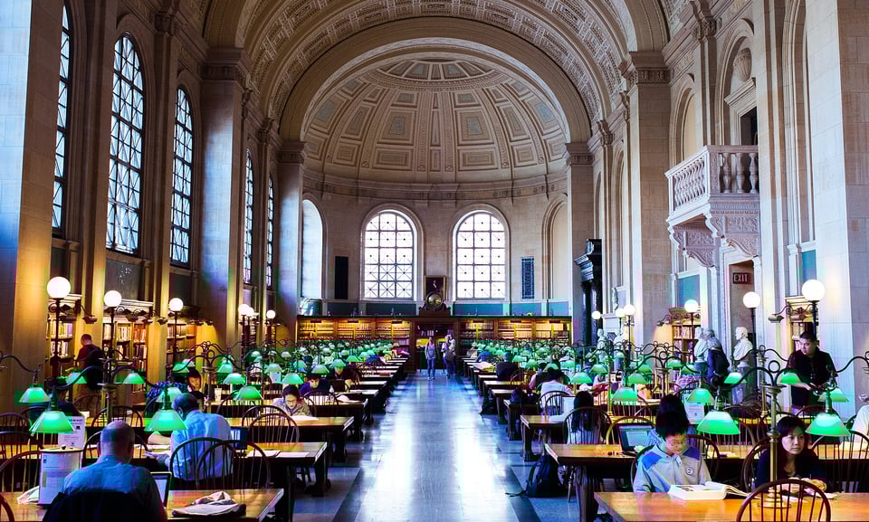 The reading room of the Boston Public Library. A huge, vaulted space done in elegant 19th-century style, with dozens of rows of desks, each with two green lamps hanging above them and many with readers seated at the desk.