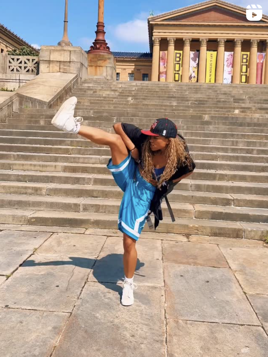 I'm dancing on the steps of the Philadelphia Museum of Art, mid-move with one leg lifted high and my arms strong and expressive. I'm wearing a black cap, a sheer black shirt, a blue sports bra, and bright blue basketball shorts with white trim, paired with white sneakers. The iconic museum columns and banners are in the background under a bright blue sky.