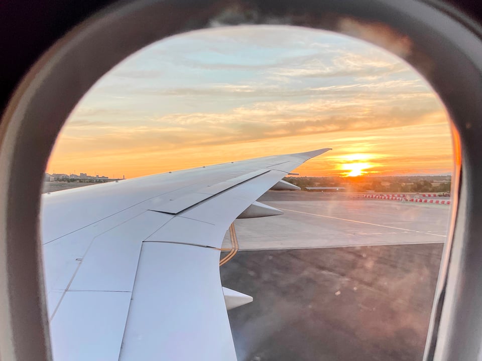 Looking out an airplane window, the wing stretches over the cement. The orange sun kisses the horizon.
