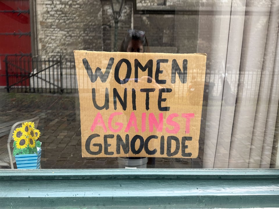 A cardboard sign is in a window and it reads "Women Unite Against Genocide".