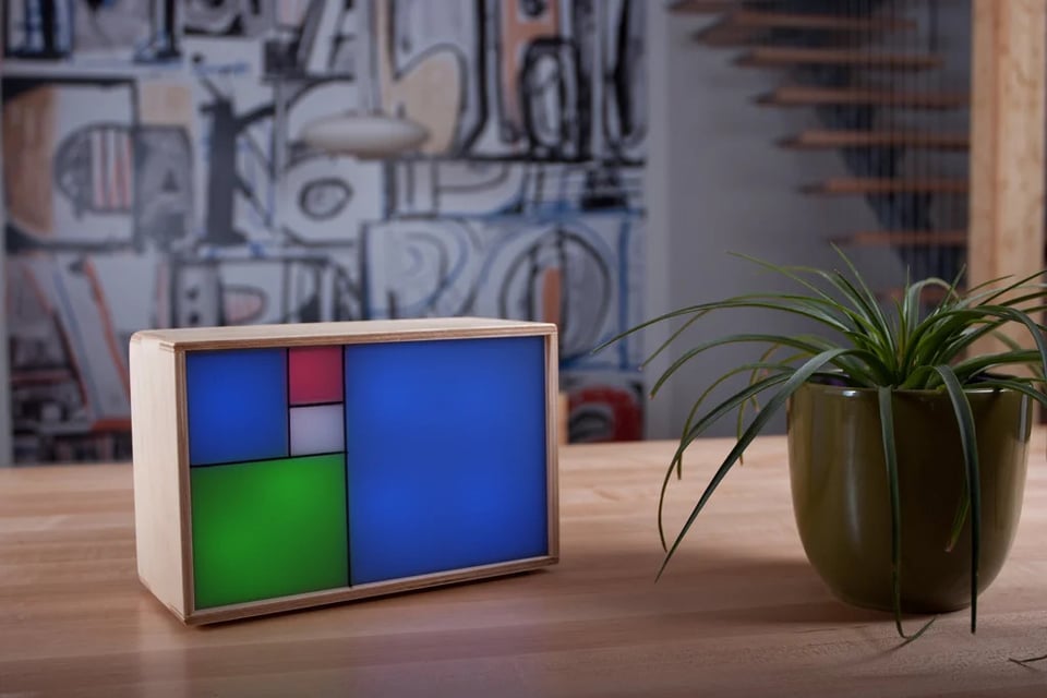 A photo of the Fibonacci clock sitting on a wooden counter with a spider plant next to it, and, in the far background, what looks like some sort of graffiti painting. The clock face is divided into six squares - one large blue one, a slightly smaller green one, a slightly smaller blue, one, and two very small ones colored red and white. It looks a little bit like a Mondrian painting.
