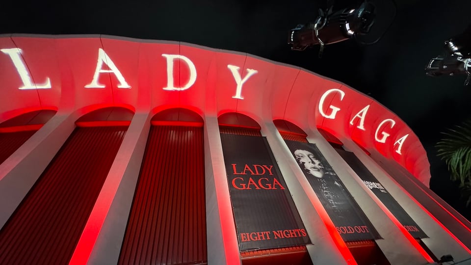 The outside of the Kia Forum is lit up in red and LADY GAGA spelled out in white all caps on the top rim. There are black banners hanging that read LADY GAGA EIGHT NIGHTS and a photo of Lady Gaga that says SOLD OUT underneath.