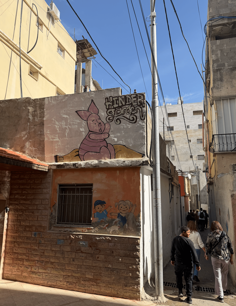Photograph of a narrow urban alley featuring a mural of Piglet from Winnie the Pooh with the English word ‘KINDER garden’ above it. The mural is painted on a building corner, with additional smaller cartoon characters below a barred window, while several people walk along the alley under a blue sky. Many electrical wires run above the buildings