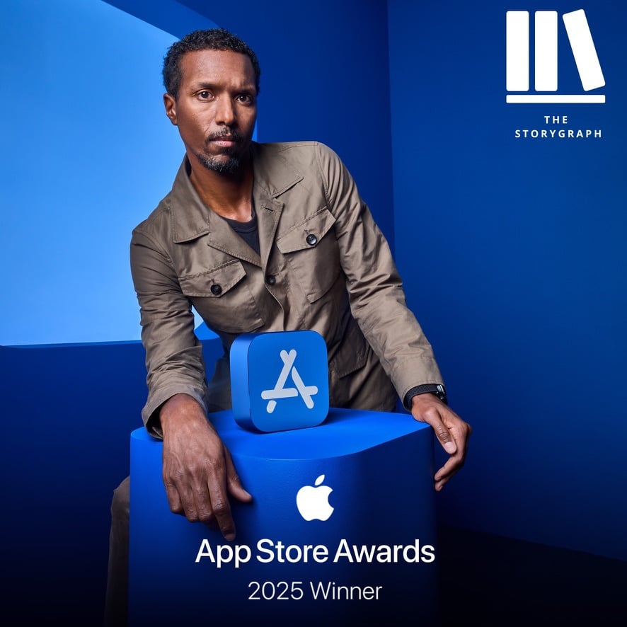 A man stands in a vivid blue studio, leaning over a blue pedestal that holds a blue App Store Awards trophy marked with the white App Store icon. He wears a tan jacket and looks directly at the camera. The white Apple logo and text “App Store Awards — 2025 Winner” appear on the front of the pedestal. In the top-right corner, The StoryGraph logo is displayed.