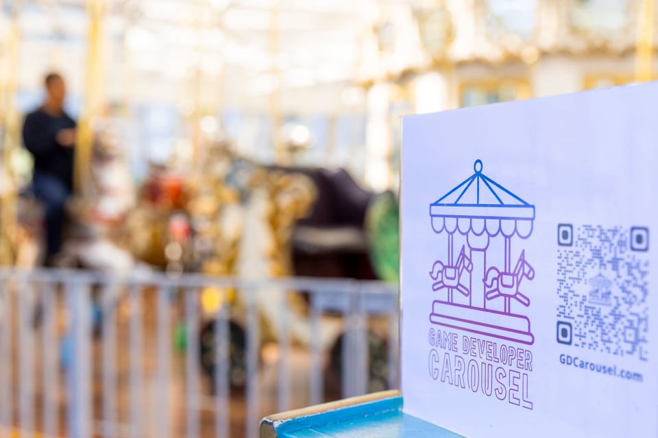 A sign for Game Developer Carousel in the foreground. The LeRoy King Carousel in the background, out of focus.