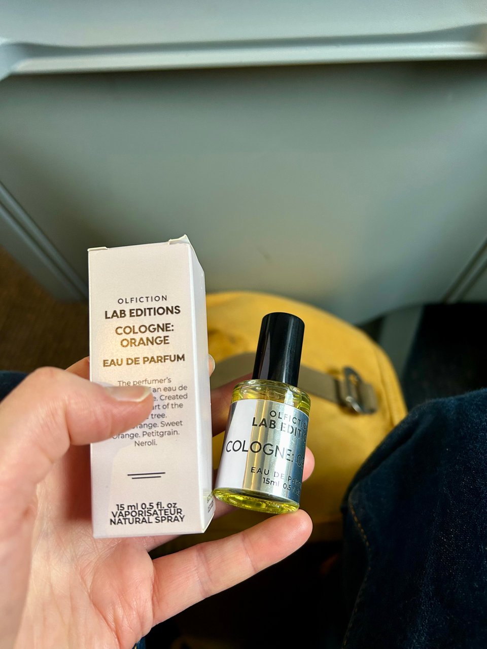 A small glass bottle of perfume with a silver label is being held with its box by a white person's hand. There's a train seat back behind and a yellow backpack below. Image by Rowan Ambrose.