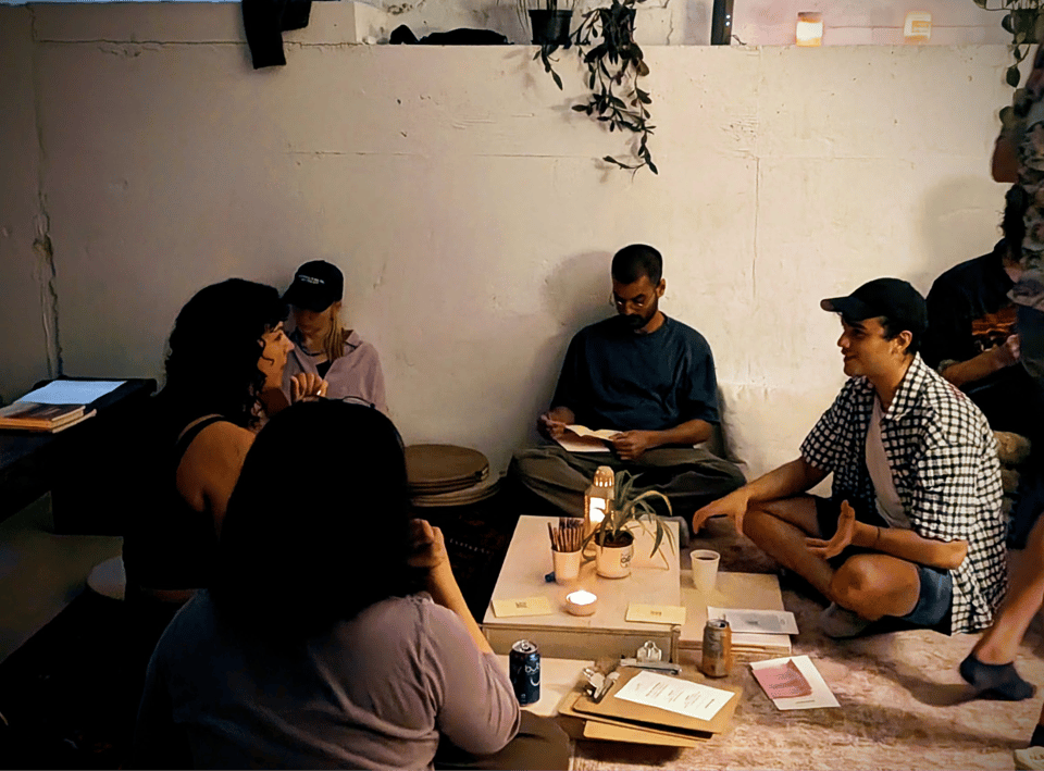 Five people sit cross legged on the floor around a low table with plants and candles on it, engaged in conversation.
