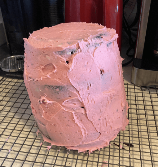 A tall, pink-iced cake sits on a cooling rack.