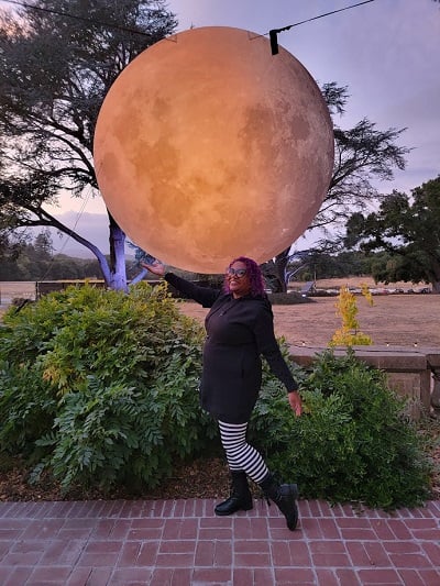 Patricia in a black dress and striped tights standing under a large moon art piece