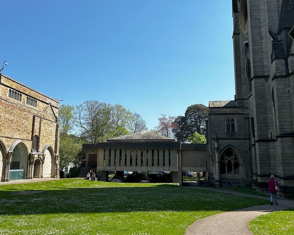 A low single storey concrete building with lots of tall slit windows rests on some pillars beneath a blue sky. To the left is a mishmash of archways into a square building. To the right, and casting shadows, is the northern side of a gothic cathedral.